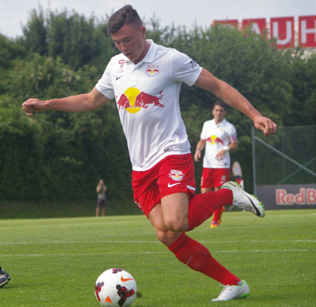 Ein professioneller Fußballspieler, der einen Fußball auf einem grünen Feld mit Bäumen und einem klaren blauen Himmel im Hintergrund kickt, mit einer Tafel mit der Aufschrift "RB Leipzig" im Hintergrund.