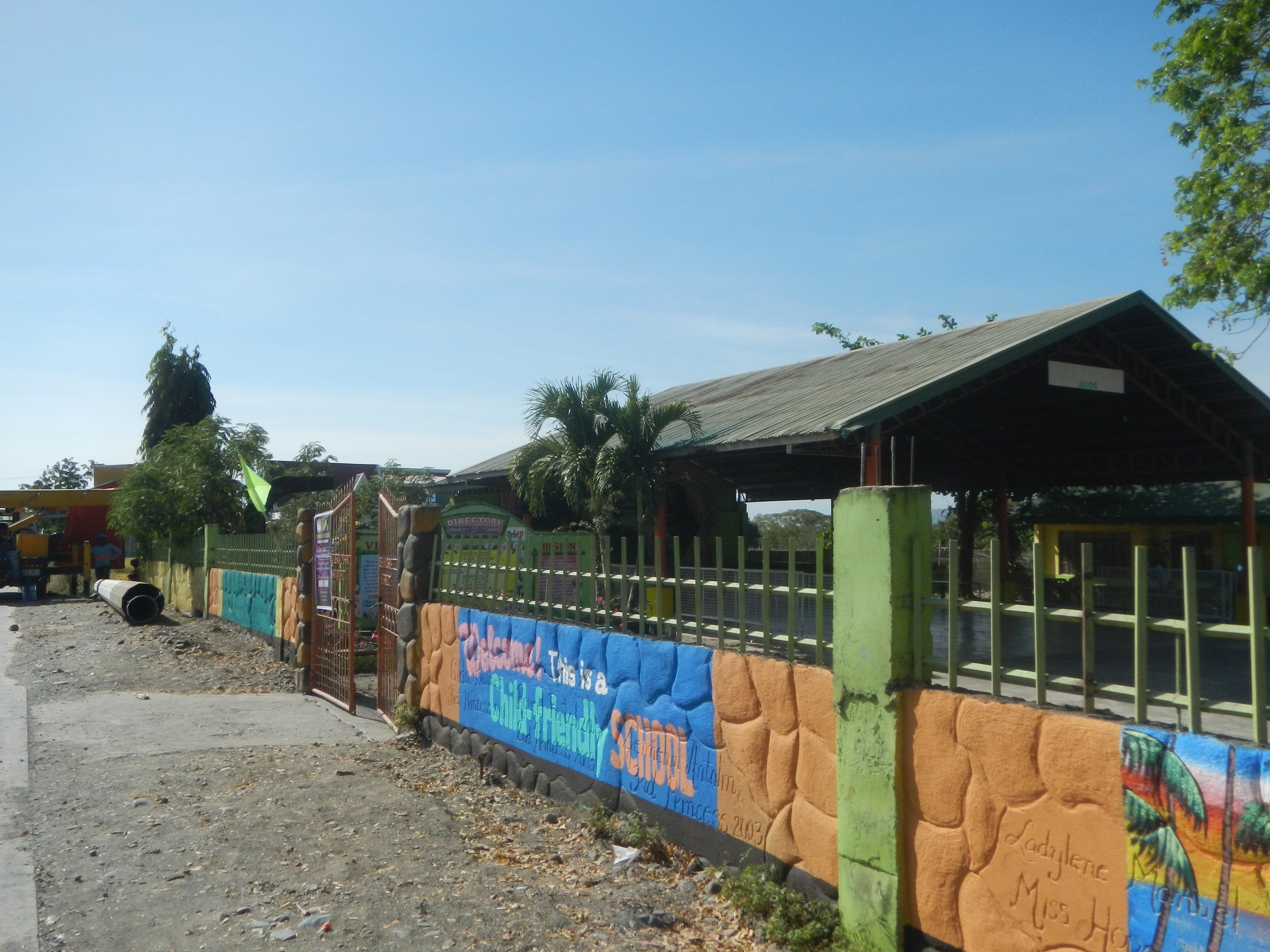 Schulgebäude mit Wandgemälde, umgeben von einem Zaun, einem Schuppen, Bäumen, Fahrzeugen, Strommasten und einem bewölkten Himmel.