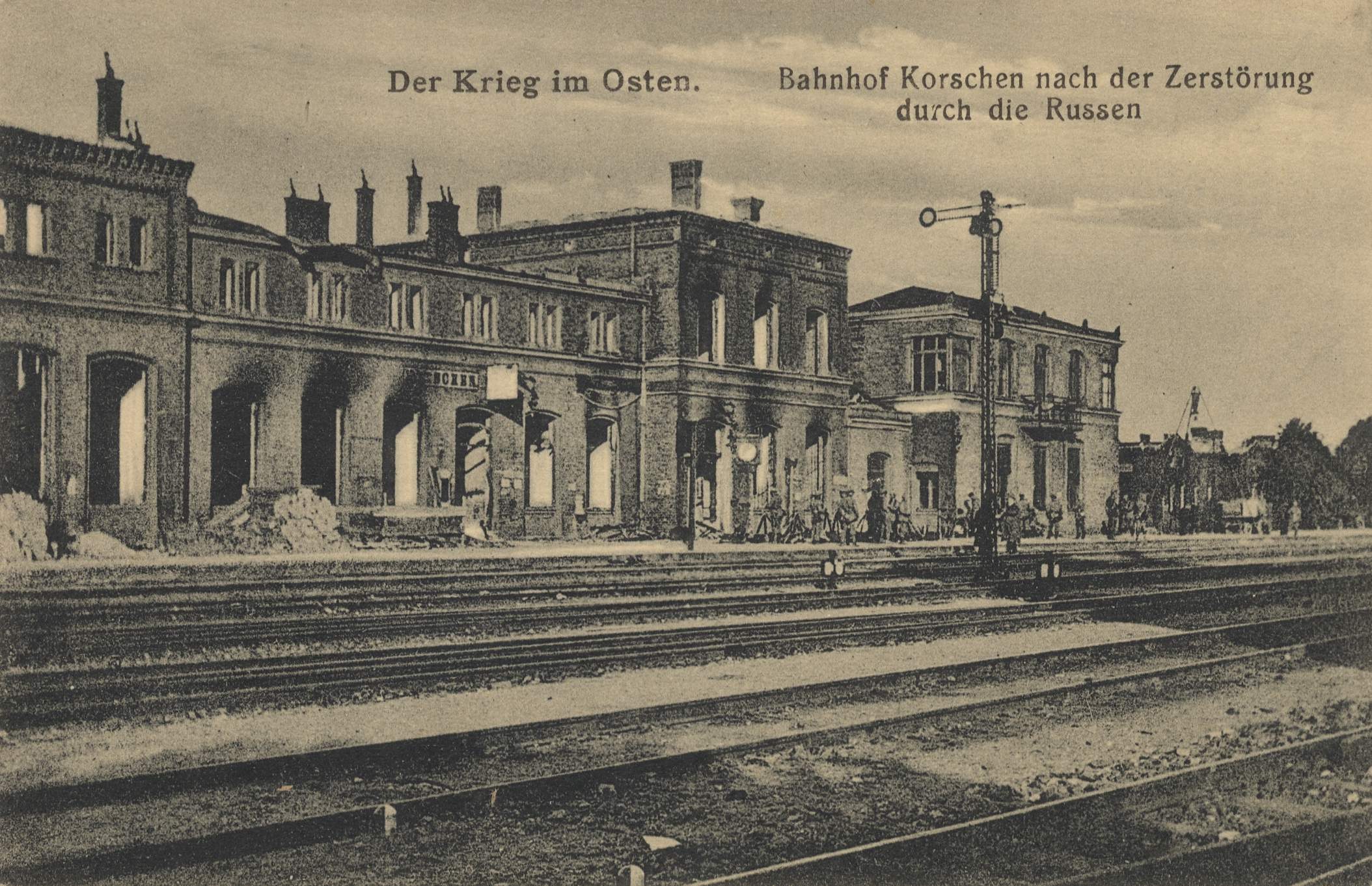 Schwarz-weiß-Fotografie eines Bahnhofs mit dem Titel "Bahnhof Korschen in Ostern, Deutschland" mit Bahnschienen, Gebäuden, Pfählen, Menschen, Bäumen und einem Himmel im Hintergrund.