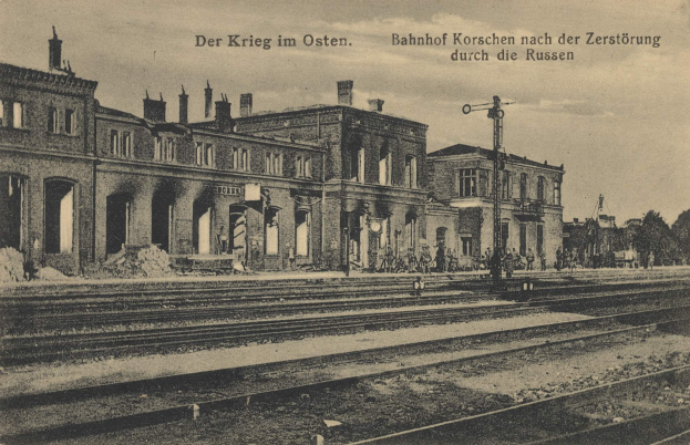 Schwarz-weiß-Fotografie eines Bahnhofs mit dem Titel "Bahnhof Korschen in Ostern, Deutschland" mit Bahnschienen, Gebäuden, Pfählen, Menschen, Bäumen und einem Himmel im Hintergrund.