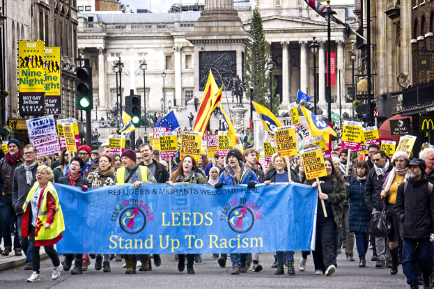 Gruppe von Menschen marschiert mit einem 'Stand Up to Racism'-Schild die Straße entlang, mit Gebäuden, Bäumen, Laternen, Ampeln und einer Statue im Hintergrund bei bewölktem Himmel.