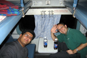 Ein Abteil mit zwei Liegen, jeweils mit einer Decke und einem Kissen, sowie einem Tresen mit Flaschen; zwei Personen mit Brille sitzen, und es gibt ein Fenster mit einem Vorhang, sowie sichtbaren Text und Zahlen.