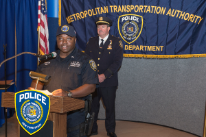 Zwei Polizisten stehen vor einem Rednerpult, einer hält Papiere und spricht in ein Mikrofon, mit einer Fahne, einem Banner und einem Tisch in der Nähe und einer Tafel mit Symbolen und Text im Hintergrund.