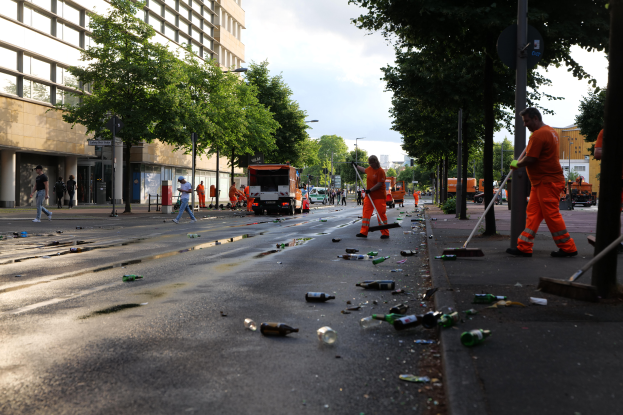 Eine Gruppe von Menschen in orangen Uniformen sammelt Müll am Straßenrand, mit Flaschen und anderem Unrat drumherum, Bäume, Pfähle, Bretter und Fahrzeuge in der Nähe und Gebäude mit Fenstern im Hintergrund bei einem bewölkten Himmel.