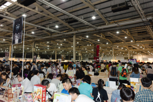 Eine große Gruppe von Menschen umringt Tische voller Bücher, Kisten und anderen Gegenständen auf einem Buchmesse, mit Bannern und Lichtern darüber.