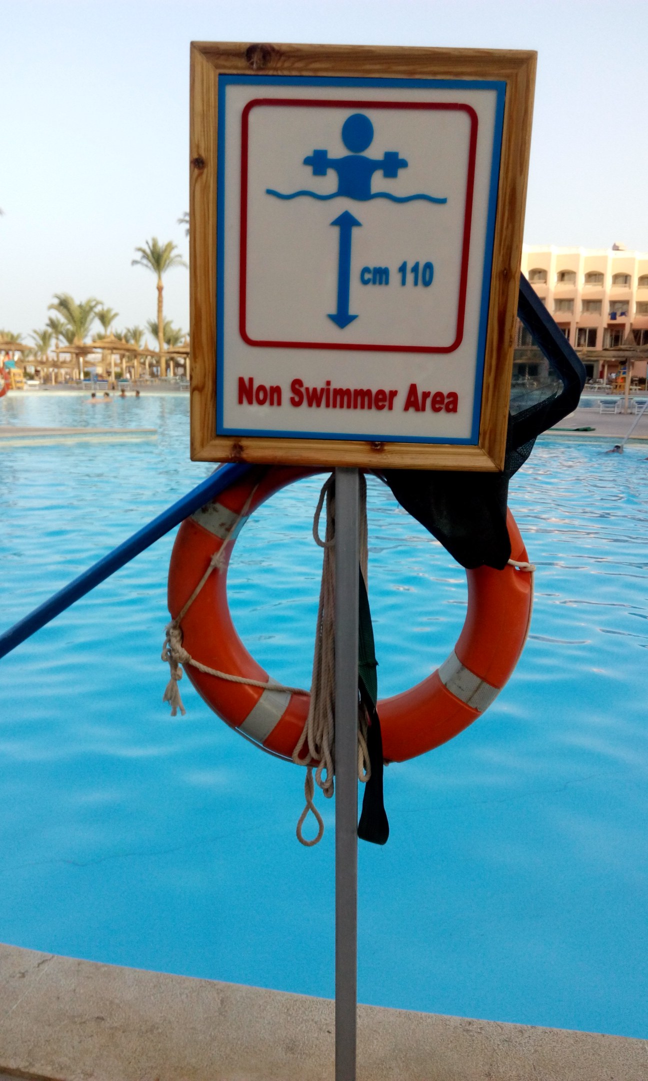 Ein Schwimmbad mit einem "Kein Schwimmer-Bereich"-Schild und einem Rettungsring an einem Pfahl, umgeben von Bäumen, Gebäuden und einem klaren blauen Himmel.
