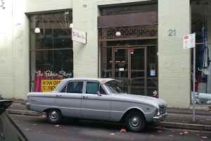 Ein silberfarbenes Auto parkt vor einem Gebäude mit mehreren Türen, einem Schild an einem Pfahl und einer Straße im Hintergrund.
