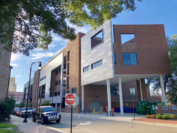 Ein großes Gebäude mit zahlreichen Fenstern, identifiziert als City Hall Apartments, umgeben von Straßenmasten, Fahrzeugen, Schildern, Verkehrskegeln, Mülltonnen, Büschen und Bäumen unter einem bewölkten Himmel.