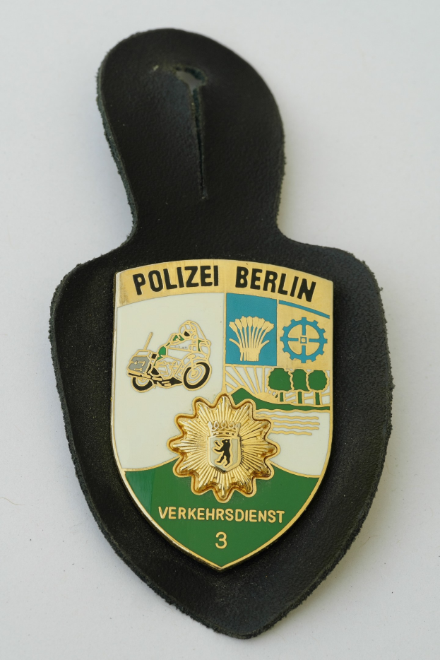 Ein schildförmiges Polizeibadgeb mit "Polizei Berlin" in schwarzer fetter Schrift, das ein weißes Stern auf einem blauen Hintergrund und einen weißen Rand hat, auf einer Oberfläche platziert.
