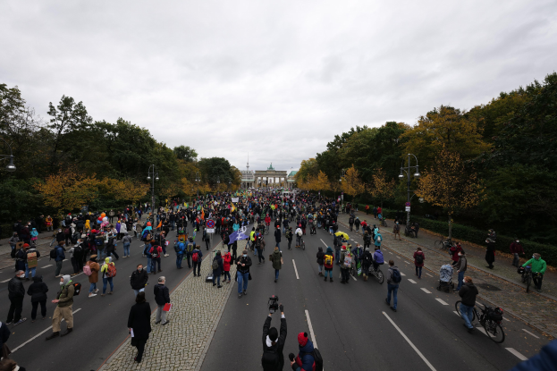 Eine große Gruppe von Menschen geht auf einer von Bäumen gesäumten Straße mit Laternenmasten, die Kameras halten, während sie an einer Protestdemo in Berlin teilnehmen.