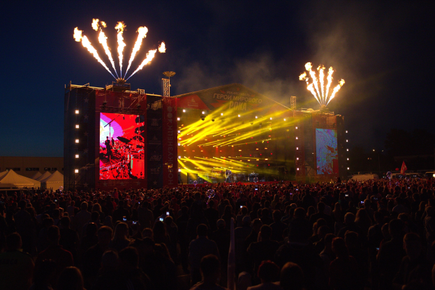 Eine große Menschenmenge bei einem Musikfestival schaut auf einer hell erleuchteten Bühne Performern zu, mit Zelten links und Feuerwerk im dunklen Himmel dahinter.