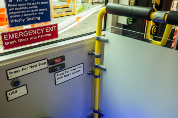 Notausgangsschild an einer Zugseite mit Tür, Griff und Glasfenster, das Straße, Pfosten und Objekte draußen zeigt.