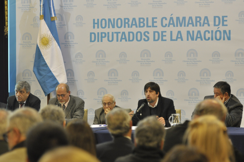 Eine Gruppe von Menschen sitzt in Reihen von Stühlen, mit einem Tisch vor der ersten Reihe, der einen Krug, Papiere und einen Stift enthält. Eine Person in der Mitte spricht in ein Mikrofon, mit einer weißen und blauen Flagge dahinter und einem weißen und blauen Hintergrund, der "ehrenwerte kamara de" anzeigt.