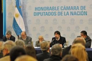 Eine Gruppe von Menschen sitzt in Reihen von Stühlen, mit einem Tisch vor der ersten Reihe, der einen Krug, Papiere und einen Stift enthält. Eine Person in der Mitte spricht in ein Mikrofon, mit einer weißen und blauen Flagge dahinter und einem weißen und blauen Hintergrund, der "ehrenwerte kamara de" anzeigt.