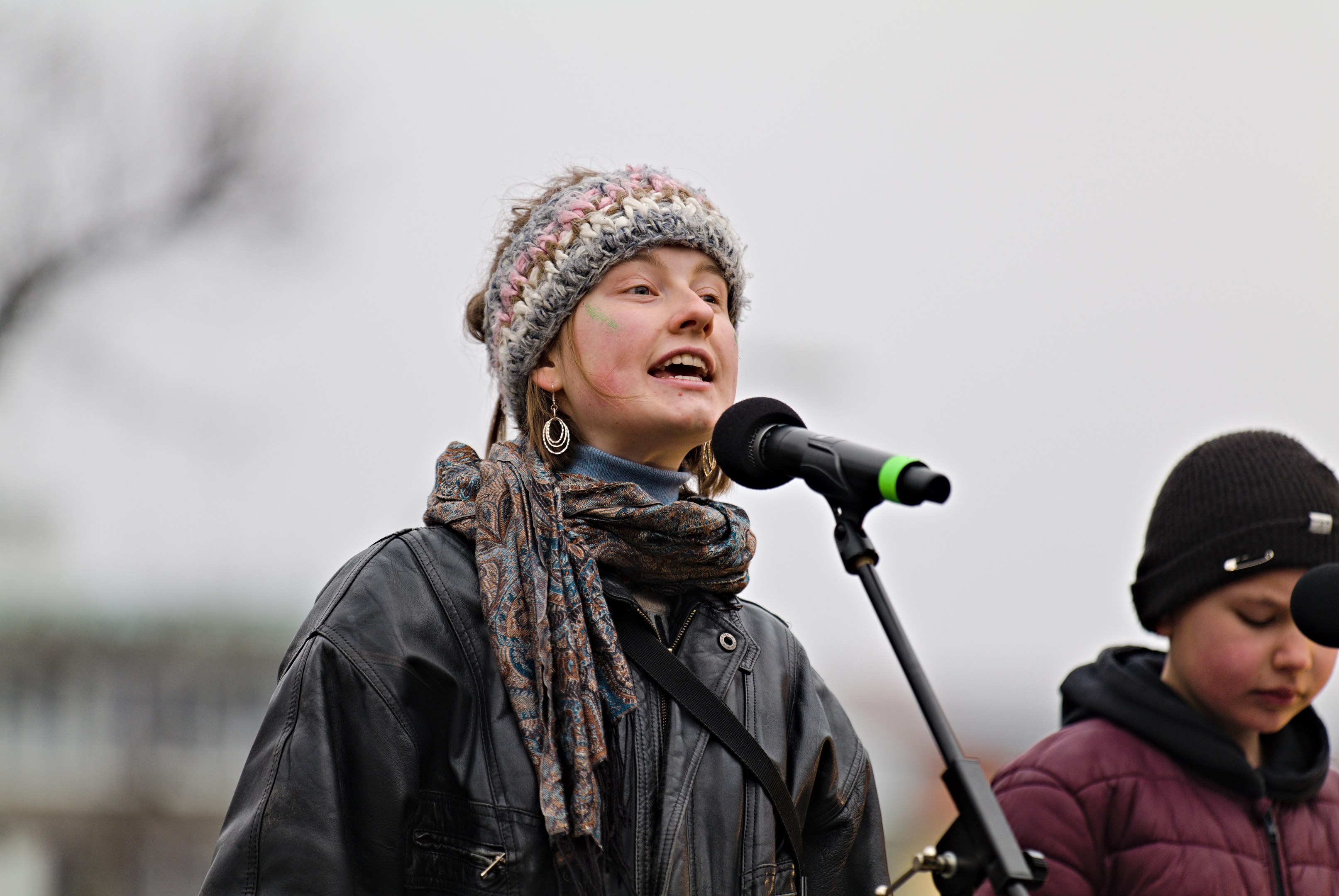 Eine Frau in einer schwarzen Jacke, einem Schal und einer Mütze singt in ein Mikrofon auf der Bühne, während eine andere Person in einer maronenfarbenen Jacke und einer schwarzen Mütze neben ihr steht und der unscharfe Hintergrund einen träumerischen Effekt erzeugt.