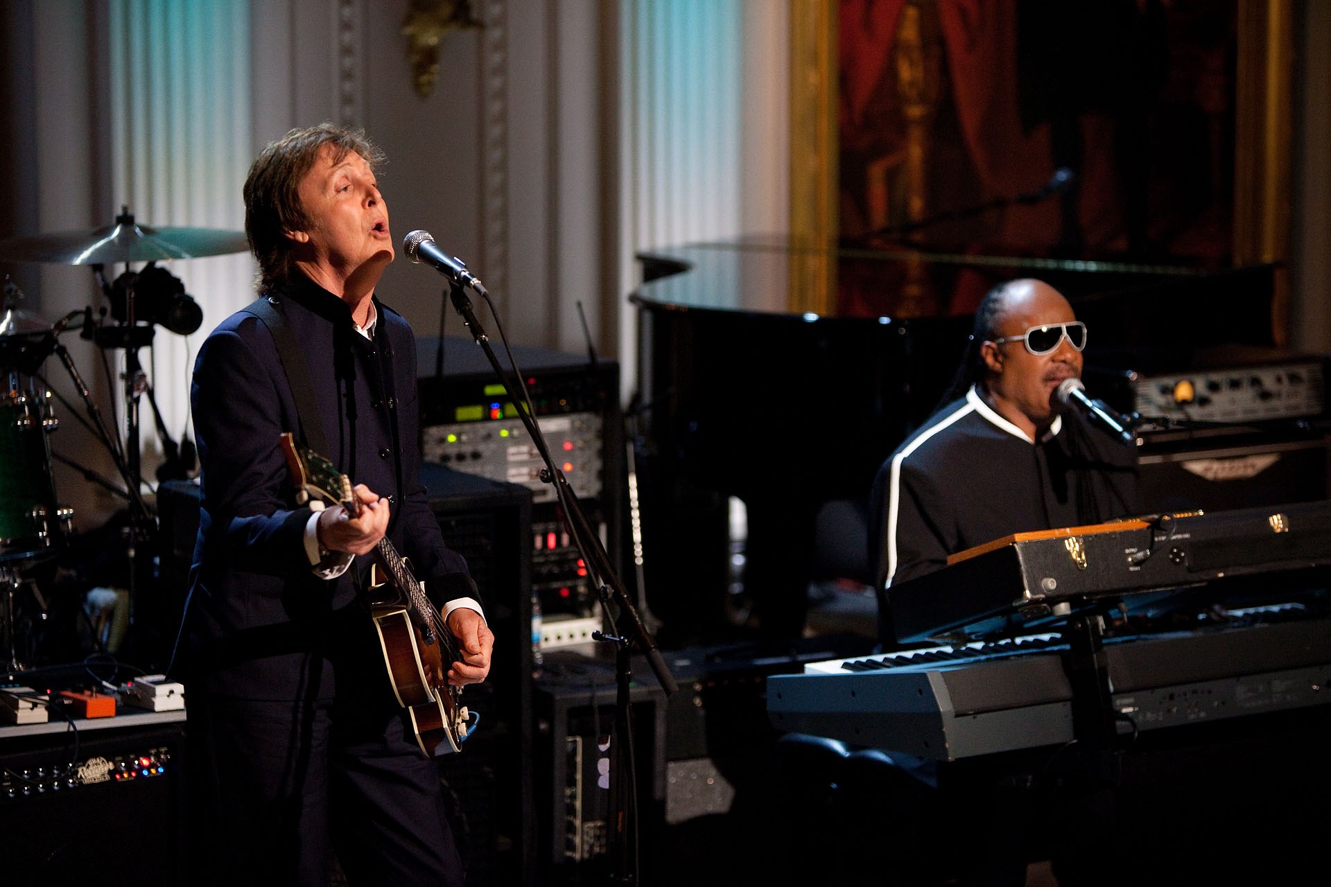 Paul McCartney spielt Gitarre und singt in ein Mikrofon, während Stevie Wonder ein Piano spielt und eine Brille trägt, auf der Bühne bei den Grammys mit Musikinstrumenten und gerahmten Kunstwerken im Hintergrund.