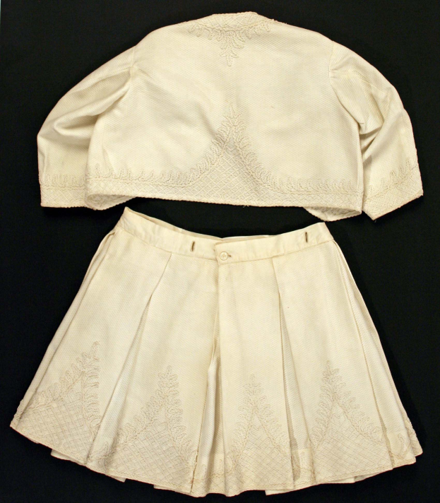 Ein hochtaillierter weißer Plisseerock und eine einfarbige weiße kurzärmlige Bluse mit rundem Halsausschnitt auf einem schwarzen Hintergrund, der Stoff zeigt eine subtile Schimmer.