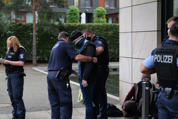 Deutsche Polizeibeamte vor einem Gebäude mit einer Frau, die ein Buch und einen Stift hält, während andere in der Nähe sitzen und stehen, mit Bäumen und Gebäuden im Hintergrund.