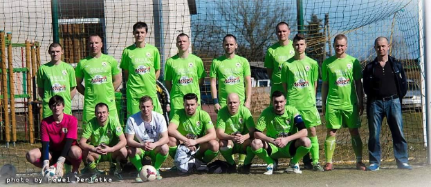 Eine Gruppe von Männern in grünen T-Shirts steht auf einem Fußballfeld in der Nähe eines Torpfostens, mit einem Ball davor und diversen Hintergrundelementen wie Bäume, Gebäude und Fahrzeuge.