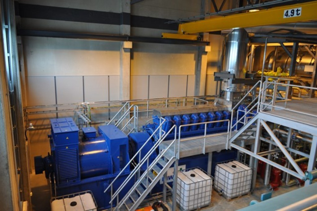 Fabrikinnere mit Trägern, einer Treppe mit Geländer, Containern, Deckenleuchten, freiliegenden Rohren und einer Wand.