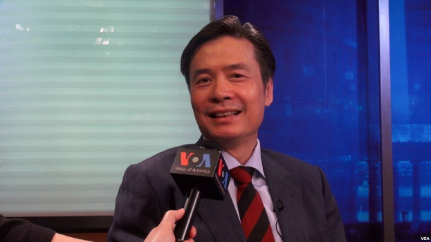 Ein Mann in Anzug und Krawatte hält ein Mikrofon und spricht vor einem Fenster und einer Wand. Links ist die Hand eines Reporters zu sehen.