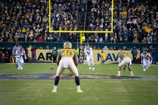 Ein Fußballspiel in vollem Gange mit Spielern in Sportkleidung und Helmen sowie Zuschauern auf den Tribünen und sichtbaren Stadionwerbung und Pfosten.