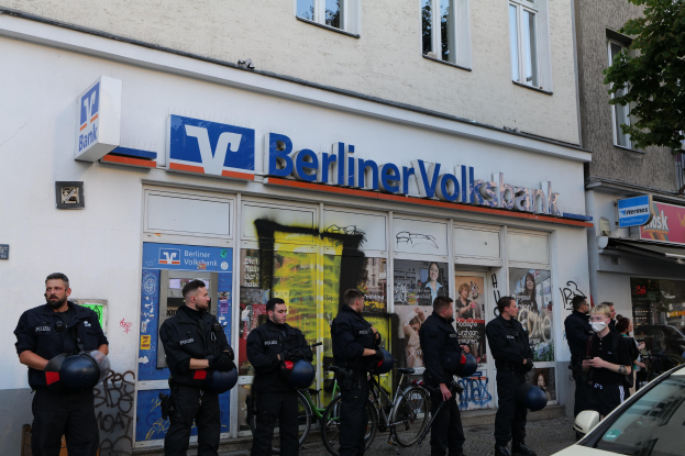 Eine Gruppe von Polizeibeamten vor einer Berliner Volksbank, einige halten Helme in den Händen, daneben ein Auto und ein Baum; im Hintergrund Gebäude mit Fenstern und Schildern.