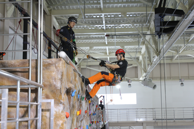 Zwei Personen in schwarzer und oranger Kleidung und Helmen klettern an einer Felswand in einer Halle, gesichert durch Seile, mit einem Metallgeländer links und einer weißen Wand mit Lampen im Hintergrund.