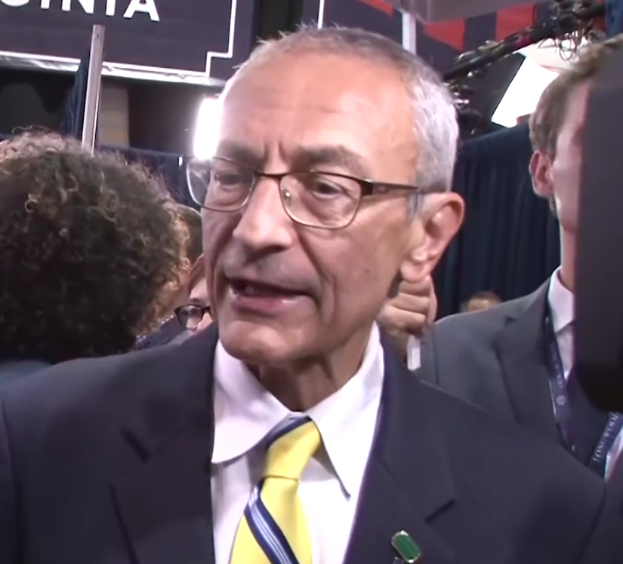 Ein Mann im Anzug und Krawatte, der eine Brille trägt, spricht zu Reportern mit einem Mikrofonständer vor ihm, umgeben von einer Gruppe von Menschen, mit Scheinwerfern und einer Tafel im Hintergrund.