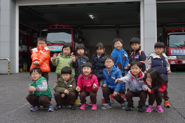 Kinder posieren vor einer Feuerwache mit Feuerwehrautos in der Garage und einem Geländer auf der linken Seite.