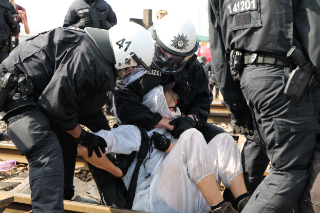 Eine Gruppe von Polizisten in Einsatzausrüstung nimmt einen Mann in einem weißen Kleid auf einem Bahngleis fest, mit anderen im Hintergrund.