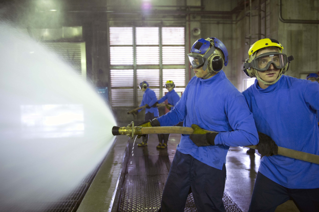 Gruppe von Männern in blauen Hemden und gelben Helmen, die an Maschinen arbeiten, wobei einer einen Schlauch hält und Wasser auf den Boden sprüht, in einer Fabrikumgebung mit Rohren, Fenstern und Lampen.