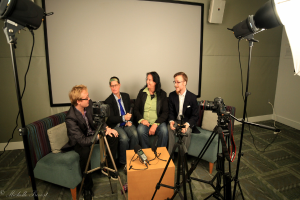 Eine Gruppe von drei Personen auf einer Couch, die ein Interview geben, mit einer Person in der Mitte, die ein Mikrofon hält, einem Recorder auf dem Tisch in der Mitte, Kameras auf beiden Seiten und einem Projektionsschirm im Hintergrund.