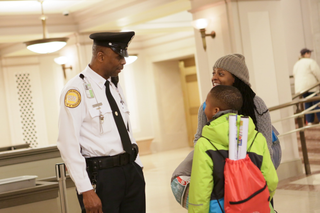 Ein Polizeibeamter spricht mit einer Frau in einer Mütze, die einen Rucksack trägt und ein Kind bei sich hat, vor einer Wand mit angebrachten Lampen und Menschen auf einer Treppe im Hintergrund.