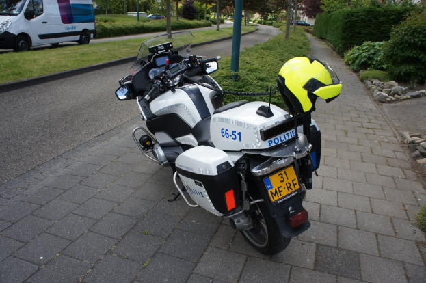 Ein auf der Straßenseite geparktes Polizeimotorrad mit einem Helm darauf, umgeben von Bäumen, Pfosten, Gebäuden, Pflanzen, Gras, Steinen und anderen Fahrzeugen im Hintergrund.