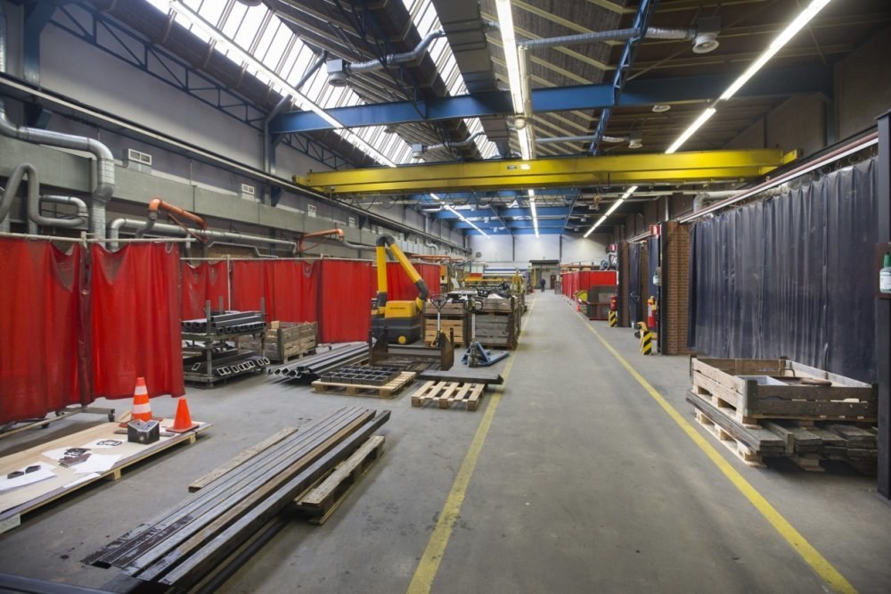 Großer Lagerraum mit Holzpaletten, Metallobjekten, Verkehrskegeln und Maschinenfahrzeug; rote Vorhänge links, Vorhangwand rechts, Deckenrohre und Lampen.