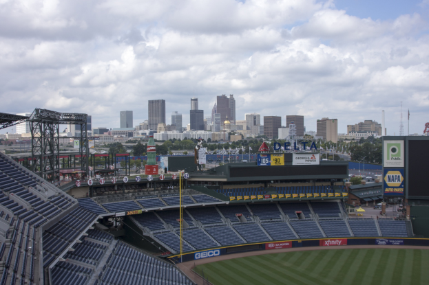 Eine wolkenverhangene Sicht auf ein Baseball-Stadion mit leeren Sitzplätzen, umgeben von einer Stadtkulisse, Rasen, Bäumen und fernen Gebäuden.