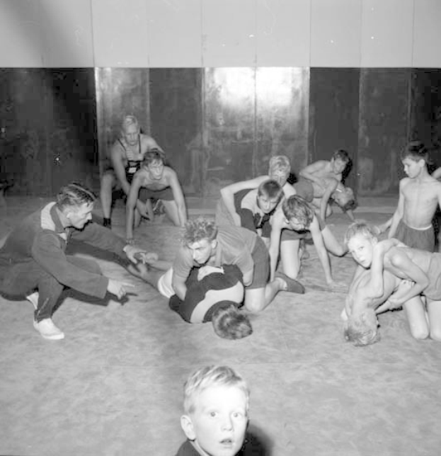 Schwarz-weißes Bild von jungen Jungs, die in einer Turnhalle räumen, mit einer Wand im Hintergrund.