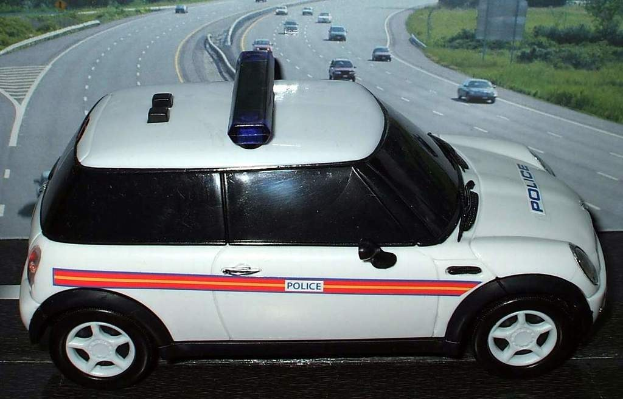 Weißer Polizeiwagen mit "Polizei" Aufschrift vor einer belebten Straße, flankiert von Gras und Bäumen außerhalb der Stadt.