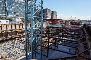 Das Bild zeigt eine Baustelle mit Gebäuden und Büros auf der linken Seite, Eisenstangen und Maschinen auf der rechten Seite und verschiedene Geräte und Kräne in der Mitte, unter einem Himmel in der oberen rechten Ecke.