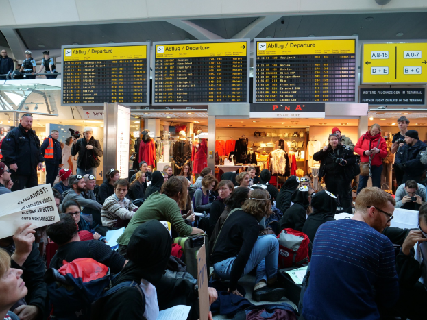 Eine große Gruppe von Menschen sitzt und steht in einem Flughafen während einer Demonstration, einige halten Taschen und Papiere, mit Schildern und Puppen im Hintergrund unter Deckenlampen.