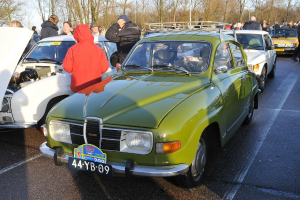 Mehrere Autos mit Menschen in der Nähe, die meisten tragen Jacken, vor einem Hintergrund aus Bäumen, Pfählen und dem Himmel.