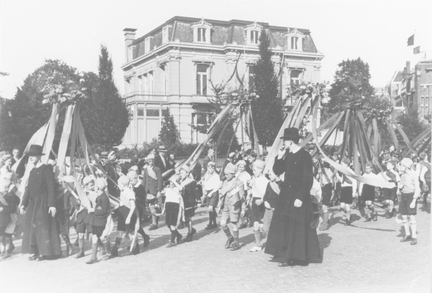 Schwarz-weißes Bild einer Gruppe von Menschen, die eine Straße entlanggehen, einige halten Fahnen, vor einem Gebäude mit Bäumen und anderen Gebäuden im Hintergrund.