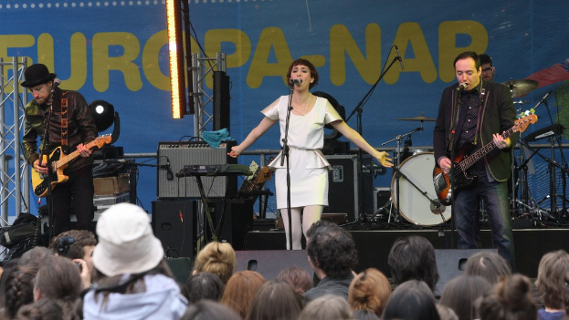 Vier Personen auf der Bühne: eine Frau in einem weißen Kleid singt in der Mitte, flankiert von zwei Männern, die Gitarren spielen, mit einer Menge unten und Lautsprechern gegen eine Wand im Hintergrund.