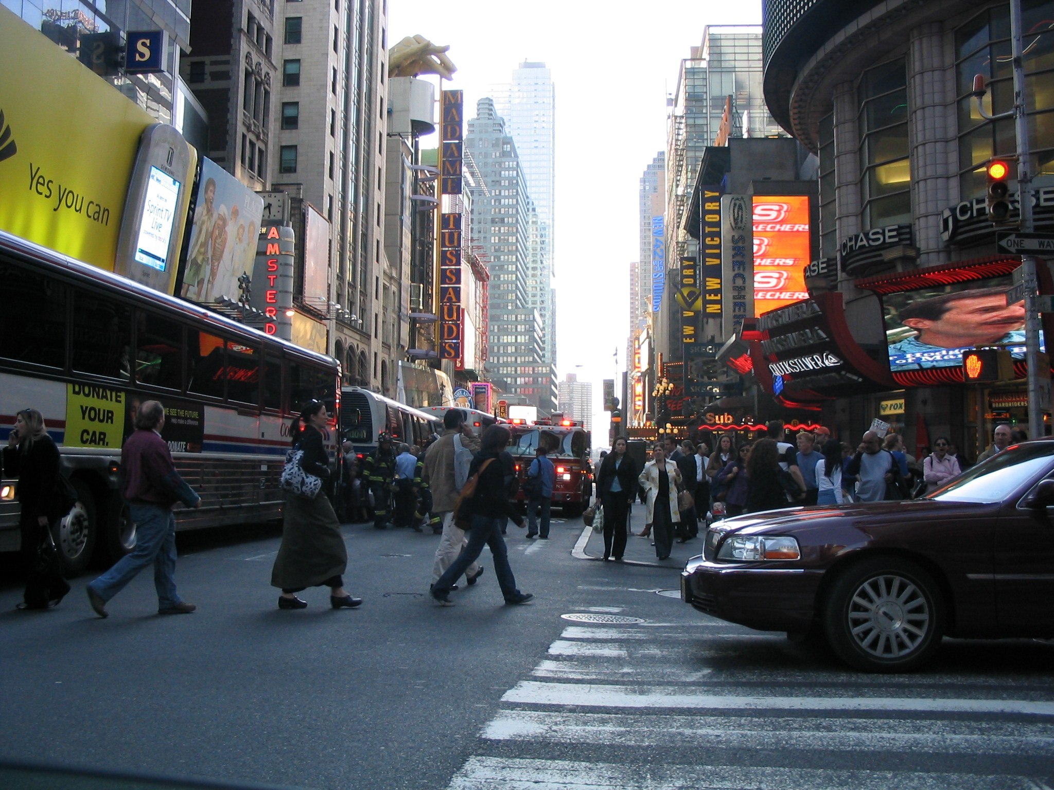 Eine belebte Stadtstraße mit Fahrzeugen, Fußgängern, die an einer Zebrastreifen überqueren, Gebäuden, Schildern, Verkehrsampeln und einem klaren blauen Himmel.