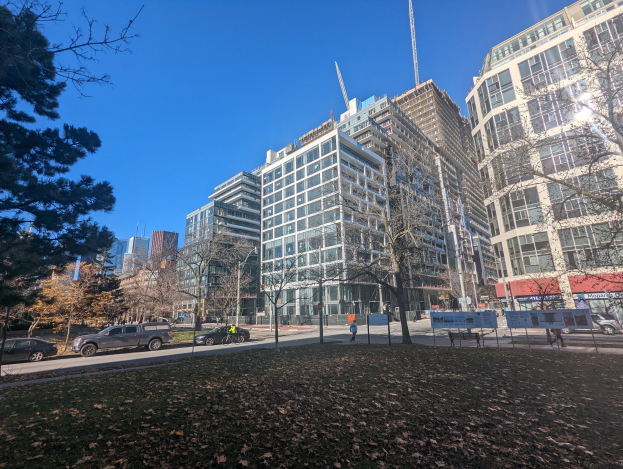 Eine Stadtstraße in Toronto mit hohen Gebäuden, fahrenden Fahrzeugen, Passanten, Bäumen und verstreuten trockenen Blättern auf dem Boden unter einem sichtbaren Himmel.