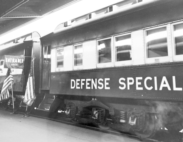Schwarz-weiß-Foto eines Zuges mit der Aufschrift "Defense Special" auf einem Bahngleis mit zwei Fahnen links und einer Decke oben.