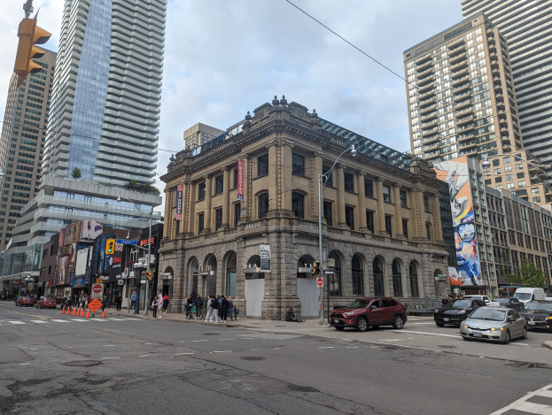 Eine belebte Stadtstraße mit hohen Gebäuden, Fahrzeugen, Fußgängern, Verkehrsinfrastruktur und Bäumen, die auf die Toronto Public Library unter einem bewölkten Himmel ausgerichtet ist.