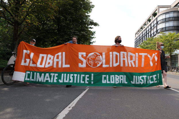 Gruppe von Menschen, die eine Straße mit einem "Globale Solidarität: Klimagerechtigkeit"-Schild entlangmarschieren, während Fahrräder entlang des Bürgersteigs geparkt sind und Gebäude im Hintergrund zu sehen sind.