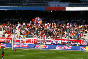 Ein Fußballspiel in einem Stadion mit Spielern auf dem Feld, ein Tor mit Netz, Banner, ein Metallzaun, ein Schild, ein Display und ein Dach mit Deckenleuchten.